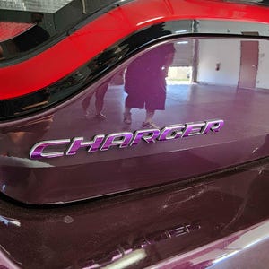 Puede incluir: Primer plano de la parte trasera de un coche granate con la palabra "CHARGER" en letras cromadas. El coche tiene un acento negro y rojo en la parte superior. Reflejos de personas y una habitación son visibles en la superficie del coche.