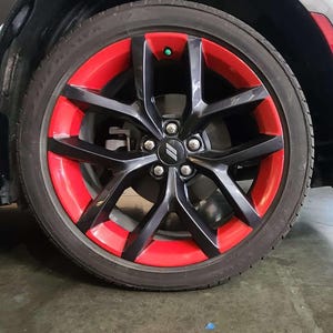 Puede incluir: Primer plano de una rueda de coche con llanta negra y detalles rojos. El neumático es negro y la rueda tiene un tapacubos plateado con un logotipo. La rueda está sobre un fondo gris.