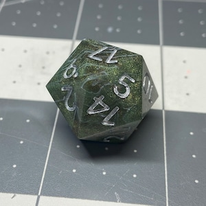 Könnte beinhalten: Ein grüner und goldener, schimmernder W20-Würfel mit silbernen Zahlen. Der Würfel ist ein Polyeder mit 20 Seiten.