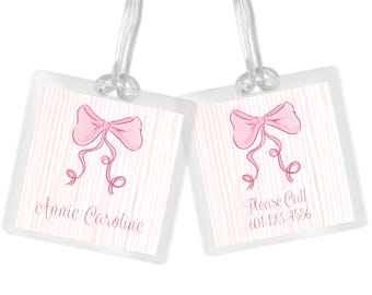 May include: Two pink and white striped luggage tags with a pink bow and the text "Annie Caroline" on one tag and "Please Call 601-123-4556" on the other tag.