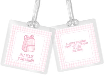 May include: Pink and white gingham luggage tag with a pink backpack illustration and the text "Ella Reese Vuncannon". The other side of the tag has the text "Ella Reese Vuncannon Thomas Street Elementary 1st Grade Mrs. Sailor".