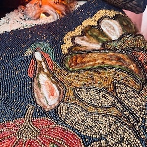 May include: A rectangular textile piece featuring intricate beadwork of a turkey and a pumpkin. The turkey is rendered in shades of brown, gold, and green beads, while the pumpkin is crafted from red and gold beads. A green beaded leaf sits below the pumpkin against a dark blue beaded background.