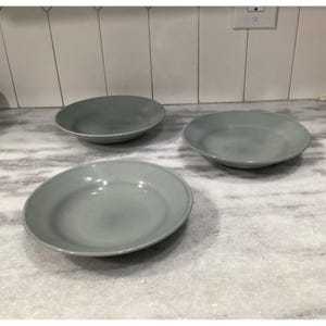 Puede incluir: Tres platos redondos de cerámica color verde salvia con un acabado mate suave y un diseño de cuenco ancho y poco profundo. Cada plato presenta un borde ligeramente elevado y un color sólido consistente en toda la superficie, dispuestos en triángulo.