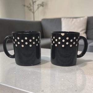 May include: Two black ceramic mugs with white polka dots arranged in a band around the top. The mugs have a curved handle and are sitting on a light gray surface. The background includes a gray couch and a white pillow.