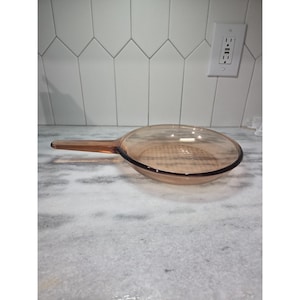 Puede incluir: Una sartén de vidrio transparente de color ámbar con un mango largo. La sartén tiene una forma redondeada con un fondo plano y un borde oscuro. El mango se extiende hacia la izquierda y la sartén está sobre una superficie de mármol.