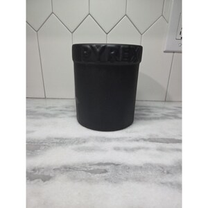 May include: A matte black Pyrex container. The word "PYREX" is embossed around the rim of the cylindrical container. The container is sitting on a white and gray marble countertop, with a white tile backsplash.
