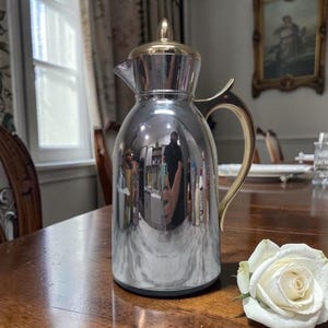 Puede incluir: Un termo plateado vintage con asa y tapa doradas. El termo es cilíndrico con un asa curva y un pico para verter. Una rosa blanca descansa sobre la mesa de madera junto al termo. El termo refleja el interior de la habitación.