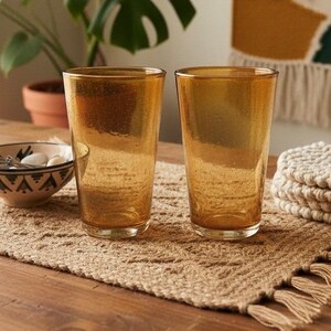 May include: Two amber-colored drinking glasses with a subtle, speckled texture throughout the glass. The tumblers have a cylindrical shape with a slightly wider rim and a smooth, clear base, reflecting light across their warm, translucent surfaces.