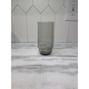 May include: A tall, gray-tinted glass tumbler. The glass has a cylindrical shape with a slightly wider base and a smooth, clear surface. The glass is empty and sits on a marble countertop.