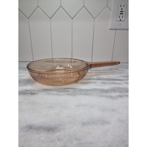 Puede incluir: Una sartén de vidrio transparente de color ámbar con un mango largo. La sartén es redonda con un fondo ligeramente curvado y un mango largo y recto. La sartén está sobre una superficie de mármol blanco y gris.