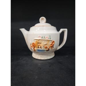 May include: Cream-colored ceramic teapot with a lid and handle. The teapot features a raised relief scene of a cozy room with a fireplace, spinning wheel, and chair. The base has a ribbed design. A small, round knob sits atop the lid.
