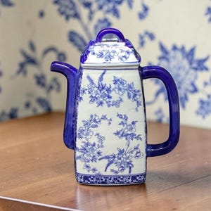 Może przedstawiać: Biało-niebieski ceramiczny imbryk o kwadratowym kształcie i pokrywce. Imbryk posiada niebieski uchwyt, dzióbek i zdobienia. Korpus zdobi kwiatowy wzór w kolorze niebieskim, tworząc klasyczny, elegancki wygląd. Imbryk stoi na drewnianej powierzchni.