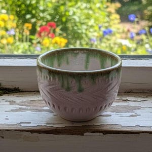 May include: A small, handmade ceramic bowl with a light pink exterior and a green rim. The bowl features a textured pattern of diagonal lines and small, evenly spaced indentations near the base. The bowl is sitting on a white windowsill.