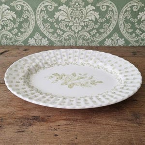 May include: A white oval serving platter featuring a scalloped edge and a repeating light green floral pattern around the rim. The center of the platter displays a detailed illustration of a rose surrounded by leaves in a soft green hue.