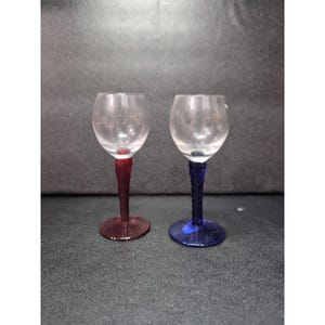 Puede incluir: Dos copas de vino pequeñas con cuencos transparentes y tallos retorcidos. Un tallo es rojo y el otro azul. Las bases de las copas son del mismo color que sus tallos. Las copas están sobre una superficie oscura.