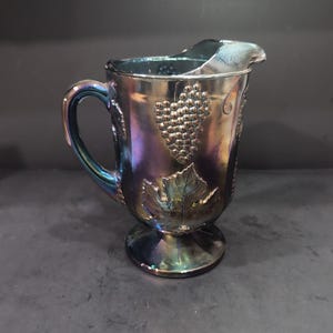May include: Iridescent glass pitcher with a handle and spout. The pitcher features embossed grape and leaf designs. The glass has a metallic sheen with colors of blue, purple, and gold. The pitcher sits on a round base.