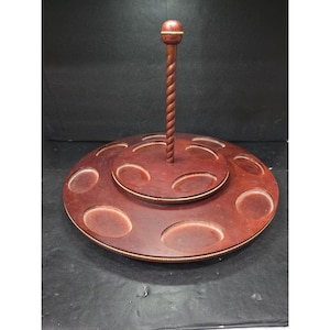 Może przedstawiać: Dwupoziomowy, drewniany stojak wystawowy z ciemnobrązowym wykończeniem. Górny poziom ma centralny, spiralny słupek zwieńczony małym, okrągłym guzikiem. Dolny poziom posiada kilka owalnych wycięć. Cienka, złota obwódka podkreśla krawędzie.