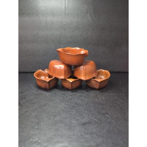 Pode incluir: Uma pilha de seis pequenas tigelas de madeira marrom. As tigelas têm uma base quadrada e uma borda arredondada em forma de pétala. As tigelas estão dispostas em forma de pirâmide, com uma tigela no topo e as outras a sustentá-la.