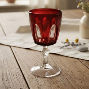 May include: A deep red glass goblet featuring a wide bowl with vertical indentations and a clear, faceted stem that transitions into a smooth, circular base. The glass has a rich, saturated crimson hue that contrasts with the transparent stem.