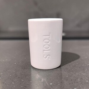 Peut inclure: Un gobelet cylindrique en céramique blanche avec le mot TOOLS embossé verticalement sur la surface avant. Le récipient présente une finition lisse et brillante, un design simple à parois droites, une base plate et un sommet ouvert.