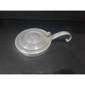 May include: A circular, silver-coloured metal pan with a lid and handle. The lid has a raised design of a planet with rings and stars. The handle is curved and attached to the side. The pan is likely for cooking, with a diameter of approximately 15 cm.