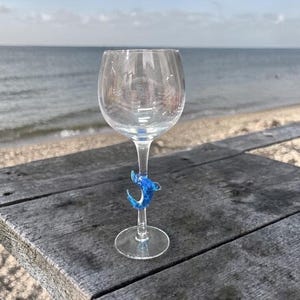 May include: A clear wine glass with a blue dolphin-shaped stem decoration. The glass sits on a weathered wooden surface with a beach and ocean in the background. The dolphin is a vibrant blue color.