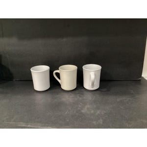 May include: Three plain white ceramic mugs arranged in a row. The mug on the left is a simple cylindrical shape, the middle mug features a curved handle, and the mug on the right has a slightly flared rim and a curved handle on its side.