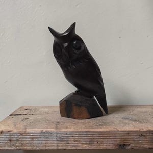 May include: A dark brown wooden owl sculpture perched on a wooden base. The owl has detailed carved feathers and a focused gaze. The sculpture is set on a rustic wooden surface, showcasing the craftsmanship.