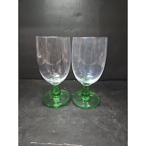 May include: Two clear glass goblets with green stems and bases. The goblets have a rounded bowl shape and are displayed against a dark background. The green bases are circular.