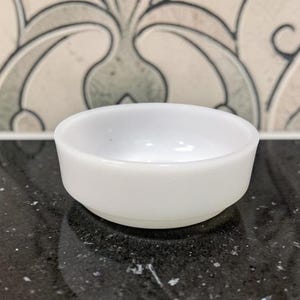 May include: A small, white, round bowl with a slightly flared rim. The bowl is made of opaque material and sits on a dark, speckled surface. The background features a decorative tile pattern.