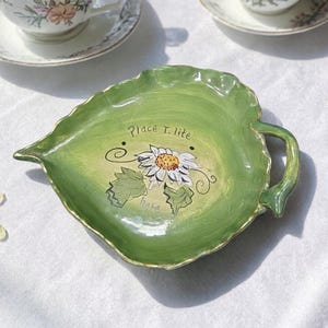 May include: A green, leaf-shaped ceramic dish with a handle. The dish features a hand-painted daisy design with the words "Place T. lite" and "have". The edges are trimmed in gold.