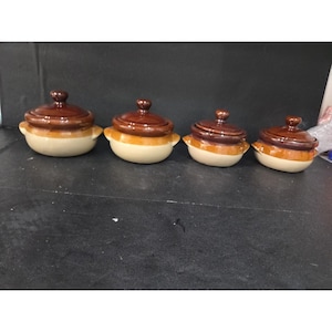 May include: Four ceramic lidded pots in a row. The pots are brown and tan with small handles on the sides. The lids are a darker brown than the base of the pots. The pots are on a dark surface.