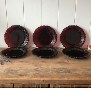 Op de afbeelding: Een set van zes vintage glazen borden. Drie zijn dieprood met een geschulpte rand en een reliëf bloemenontwerp. De andere drie zijn zwart, ovaal van vorm en hebben een vergelijkbaar reliëfpatroon. De borden zijn op een houten oppervlak geplaatst.