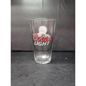 Puede incluir: Un vaso de pinta transparente con el logotipo de Coors Light. El logotipo presenta la palabra "Coors" en escritura roja sobre la palabra "LIGHT" en blanco, con un gráfico de cordillera. Un círculo blanco está encima de la cordillera.