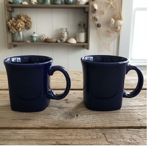Puede incluir: Dos tazas de cerámica de color azul marino oscuro a juego con un acabado liso y brillante. Cada taza presenta un asa ancha y redondeada y un borde ligeramente acampanado. Las tazas tienen forma cilíndrica y están colocadas juntas.