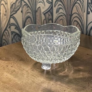 May include: A clear, vintage glass bowl with a scalloped rim and diamond-patterned sides. The bowl sits on a single, rectangular foot. The bowl is likely used for serving or display.