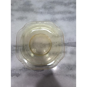 May include: A vintage, clear glass dish with a square shape and scalloped edges. The dish has a circular indentation in the center and a subtle yellow tint. The glass has a delicate, etched pattern around the rim.