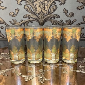 May include: Four clear drinking glasses with gold and green ornate designs. The glasses have a tall, slender shape and are arranged on a decorative tray. The gold detailing includes floral and geometric patterns, adding a vintage aesthetic.