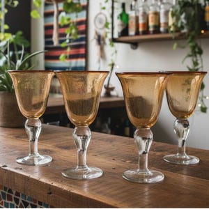 May include: A set of four stemmed glasses featuring amber-coloured bowls with a subtle speckled texture. Each glass has a clear, rounded stem and a circular base, designed with a wide, flared rim that tapers down towards the base of the bowl.