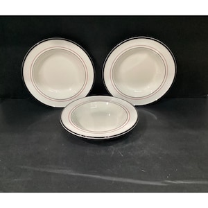May include: Three white ceramic bowls with a wide rim, each featuring a thin black outer border and a delicate red circular line accent. The bowls are arranged with two positioned in the back and one centered in the front, showcasing a smooth finish.