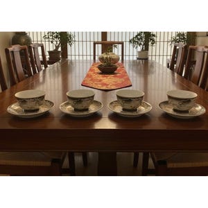 May include: Four tea cups and saucers are arranged on a dark wood table. The cups and saucers have a gold and black design. The table is set with a red runner and surrounded by wooden chairs, creating an elegant dining setting.