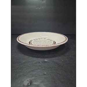 Puede incluir: Un plato de tarta de cerámica blanca con borde marrón y texto "APPLE PIE" y una receta. El plato es redondo y poco profundo, ideal para servir o hornear. El texto es de estilo vintage.