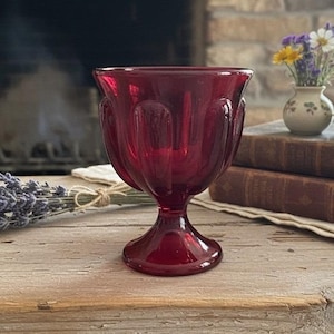 May include: A ruby red glass goblet with a decorative, fluted design. The goblet sits on a wooden surface, with a small vase of flowers and vintage books in the background. A bundle of lavender is tied with twine.
