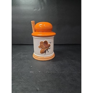 以下が含まれることがあります： オレンジ色の蓋とハンドルが付いたセラミック製の蜂蜜ポット。円筒形の本体は白で、蜂の詳細なイラストが描かれています。ベースとリムはオレンジ色で、まとまりのあるデザインになっています。キッチン装飾に適した装飾品です。