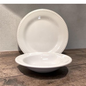 May include: Two white ceramic dishes, a dinner plate and a smaller bowl, are displayed on a wooden surface. Both pieces have a beaded edge design. The plate is in the background, and the bowl is in the foreground.