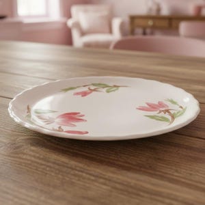 May include: A round white ceramic plate with a scalloped edge, featuring a delicate hand-painted floral pattern of pink blossoms and green leaves scattered across the surface. The plate has a smooth, glossy finish and a simple, clean design.