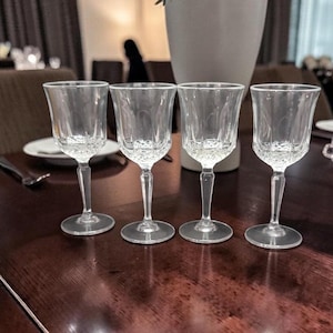 May include: Four clear crystal wine glasses with long stems and round bases are arranged on a dark wooden table. The glasses have a decorative pattern on the bowl and are empty.