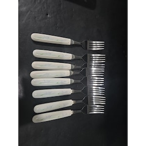 May include: A set of eight forks with white handles featuring a delicate floral design in blue and green. The tines are made of shiny silver metal, and the forks are arranged on a dark surface.