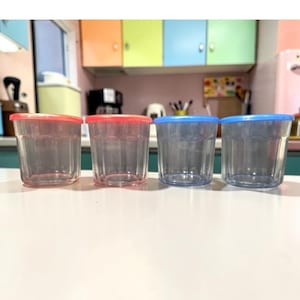 Pode incluir: Quatro copos de plástico transparente com tampas coloridas. Dois copos têm tampas vermelhas e dois têm tampas azuis. Os copos estão sobre uma superfície branca, com uma cozinha ao fundo. Os copos estão vazios.