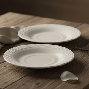 Op de afbeelding: Twee ronde witte keramische schoteltjes met een gestructureerd, kuiltjespatroon langs de buitenrand. De bordjes hebben een glad, plat midden en een licht verhoogde rand, wat een strak en neutraal ontwerp vormt voor kopjes.